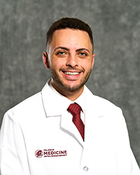 A man with short dark hair, short beard and mustache, wearing a white shirt, maroon tie and a medical white coat as he smiles at the camera.