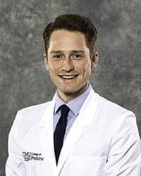 A man with brown hair wears a light colored collared shirt, dark tie and medical white coat in a headshot image.