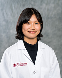 A woman with medium-length brown hair wearing a black shirt and a medical white coat as she smiles at the camera.