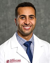 A man in a blue plaid button up, blue tie and white medical coat smiles in a head shot.