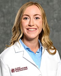 A woman with long, blonde hair and a light blue blouse wears a white coat and smiles for a headshot.