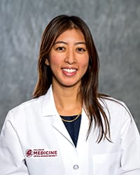 A woman with long, dark hair wears a white coat and smiles for a headshot.