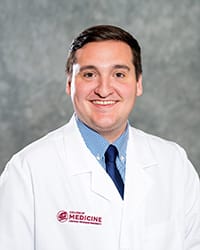 A man with brown hair wears a blue collared shirt, tie and medical white coat in a headshot image.