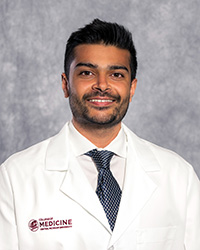 A man with short brown hair, close shaven beard and mustache wearing a white shirt, blue tie and a medical white coat as he smiles at the camera.