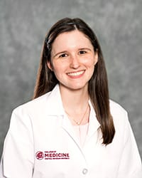 A woman with long, dark hair and a white blouse wears a white coat and smiles for a headshot.