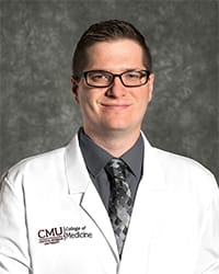 A man with brown hair wears a grey collared shirt, tie and medical white coat in a headshot image.