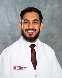 A man wearing a white lab coat, white shirt and maroon glasses as he smiles at the camera.