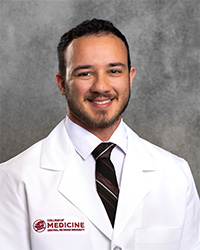 A smiling male medical student in a white medical coat stands against a gray textured background. He has dark hair, dark facial hair and wears a black tie with white stripe. The coat has a college logo embroidered in maroon.