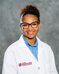 A woman wearing a white lab coat, blue shirt and glasses as she smiles at the camera.