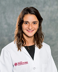 A woman with long, dark hair and a black blouse wears a medical white coat and smiles for a headshot.