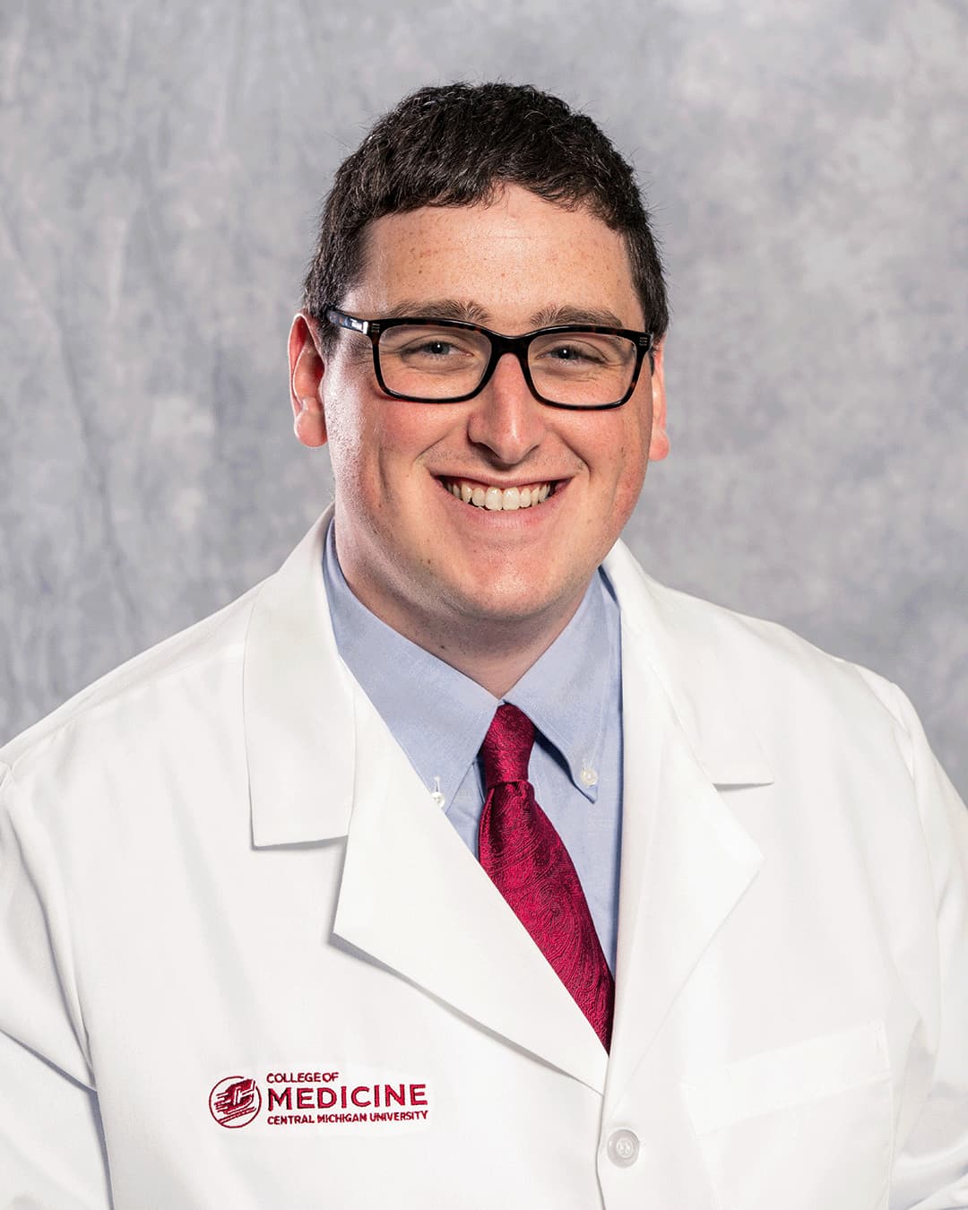 A male CMU College of Medicine medical student with rectangular black glasses is wearing his white coat as he smiles for the camera.