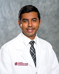 A man with short, black hair, white collared shirt and dark plaid tie wears a medical white coat and smiles for a headshot.