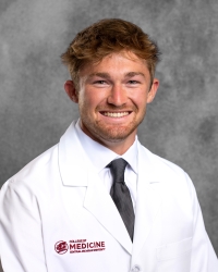 Goodwin, Zachary_student_200x250 A man with short, curly hair and facial hair is wearing a white medical coat with
