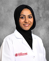 A female medical student wearing a black hijab and a white lab coat with a maroon college logo. She is smiling against a gray mottled background.