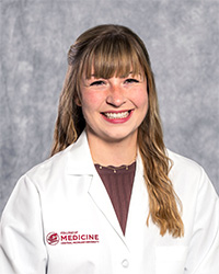 A woman with long, blond hair and a brown blouse wears a medical white coat and smiles for a headshot.