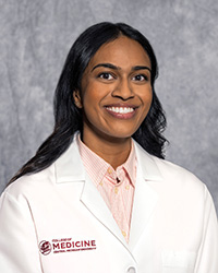 A woman with long, dark hair and a pink blouse wears a medical white coat and smiles for a headshot.