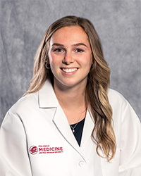 A woman with long, blond hair and a black blouse wears a medical white coat and smiles for a headshot.