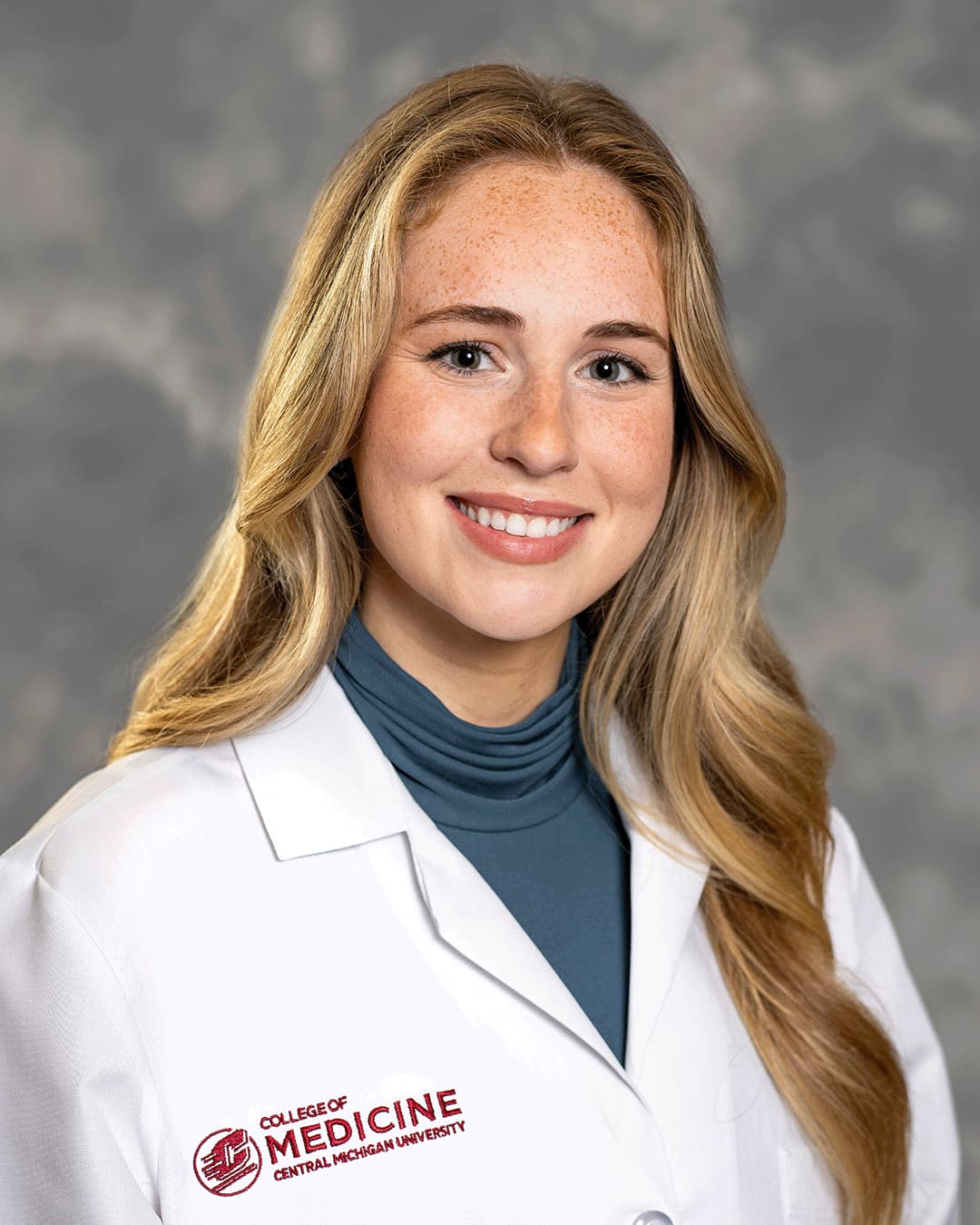A female CMU College of Medicine medical student with blonde hair is wearing her white coat as she smiles for the camera.