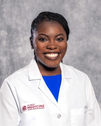 Metuge, Lenora_student_200x250 A woman is smiling, wearing a white medical coat over a bright blue top. The lab coat has a maroon logo and the words