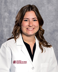 A woman with long, dark hair and a black blouse wears a medical white coat and smiles for a headshot.