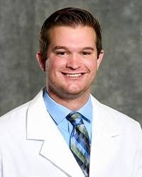 A man with short, dark hair wearing a blue collared shirt and blue plaid tie wears a medical white coat and smiles for a headshot.