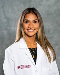 A woman with long blond brown hair wearing a blue shirt and a medical white coat as she smiles at the camera.