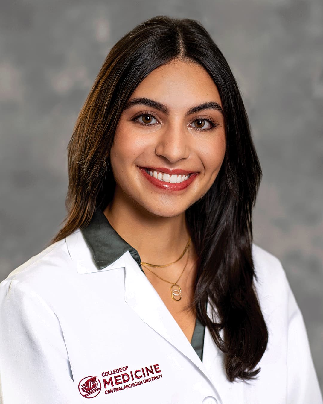 A female CMU College of Medicine medical student with long brown hair and a gold necklace wearing her white coat as she smiles for the camera.