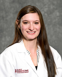 A woman with long, dark hair and a black blouse wears a medical white coat and smiles for a headshot.