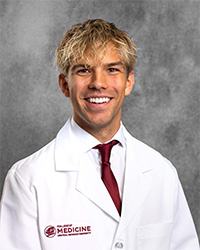 A smiling male medical student in a white medical coat stands against a gray textured background. He has light blond hair and wears a maroon tie. The coat has a college logo embroidered in maroon..