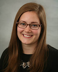 A professional headshot of Alexa Shepherd in dark attire against a dark gray background.