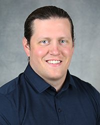 A professional headshot of Ryan Guthrie in dark blue attire against a gray background.