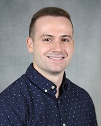 A professional headshot of Stephen Holihan in dark blue attire against a gray background.
