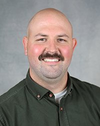 A professional headshot of Caleb Morris in dark green attire against a gray background.