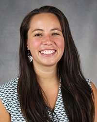 A professional headshot of Sara Schambach in light attire against a gray background.
