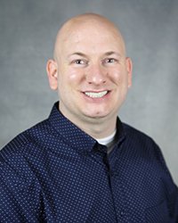 A professional headshot of Eric Dax Spencer in dark blue attire against a gray background.