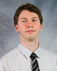 Headshot of Nicholas Lettner-Knowlton wearing a light gray dress shirt and a black and grey striped tie.