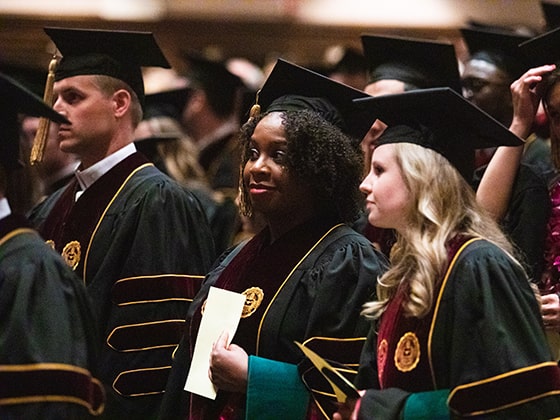 A young woman holds a paper to her chest and smiles while wearing a cap and gown and standing with her fellow graduates.