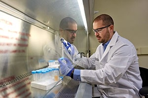 A man wearing a white lab coat, blue gloves, and safety glasses works with test tubes.