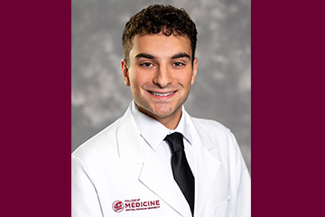 A man with dark curly hair wearing a white doctor's coat and black tie smiles at the camera.