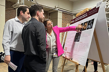 Two male students look on as a female student in a pink blazer points to her poster presentation.