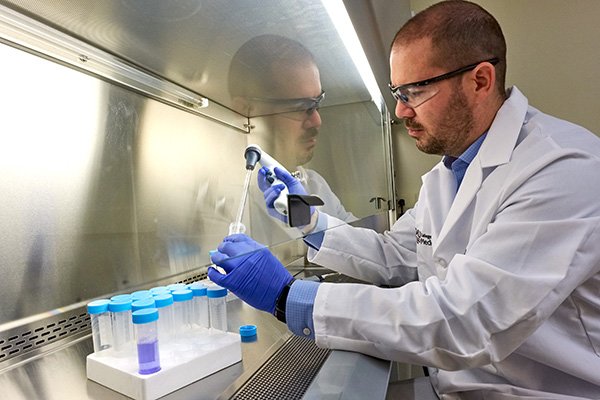 Jesse Bakke wears a white lab coat, blue gloves, and safety glasses as he works with test tubes under a fume hood.