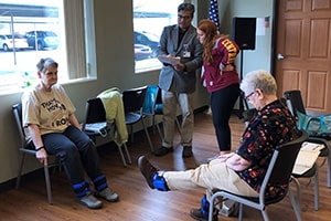 Two older women sit in chairs with cuffs strapped around their ankles, one kicking her foot up, as two people examine them and one writes down notes.