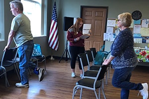 Two older adults hold the back of a chair and lift one foot up until the heel is parallel with the back of the knee.