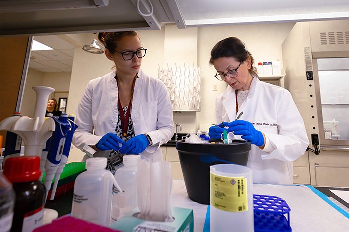 Two female medical researchers in lab coats and gloves work at a lab station. One examines a sample, while the other observes closely, surrounded by lab equipment.