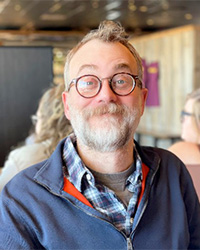 A man with short brown thinning hair, gray mustache and beard, wearing a blue plaid shirt and blue zip sweater smiles at the camera.