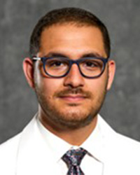 A man with short brown hair, a short beard and mustache wearing a white shirt, tie and lab coat smiles at the camera.