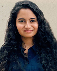 A woman with long black wavy hair wearing a blue shirt smiles at the camera.