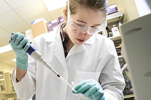A young woman wearing a white lab coat, blue gloves, and safety glasses works with a pipette and test tube in a lab during the Summer Research Scholars Program.