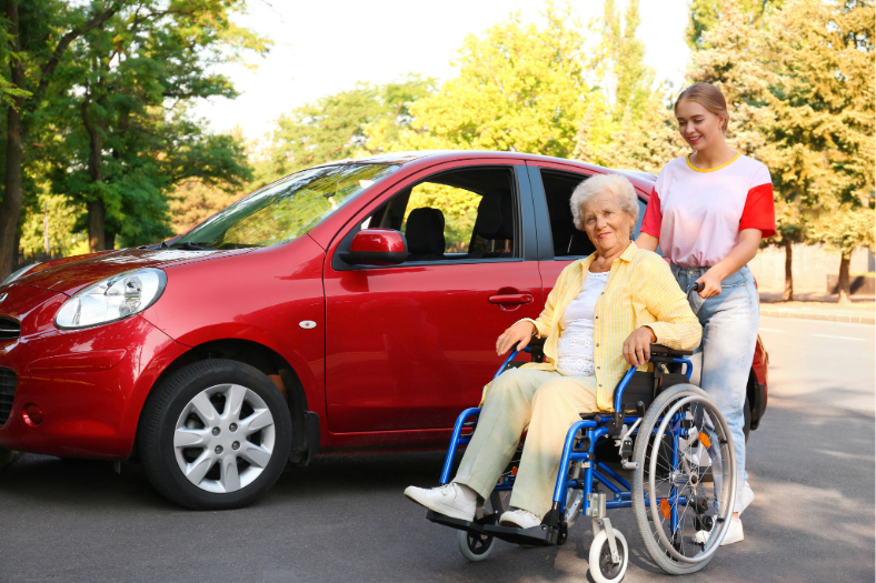 A woman pushing an elderly woman in a wheelchair next to a red SUV.
