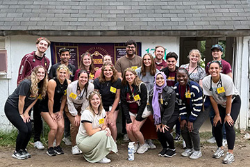 A group of medical students in flower crowns smile at the camera outside a rustic first aid clinic.